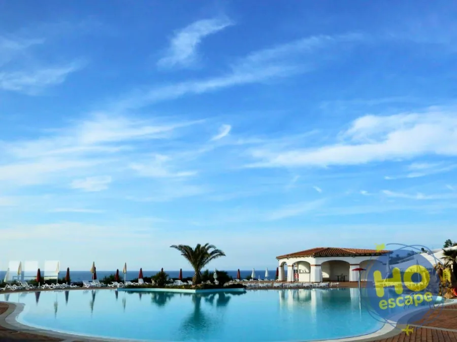 Ampia piscina con vista sul mare e cielo azzurro, ombrelloni e edificio del resort sullo sfondo