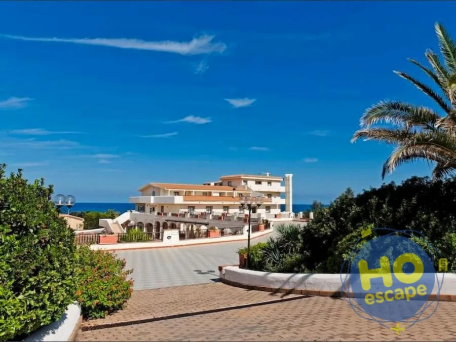 Ingresso del resort con viale pavimentato, vegetazione e edificio bianco affacciato sul mare