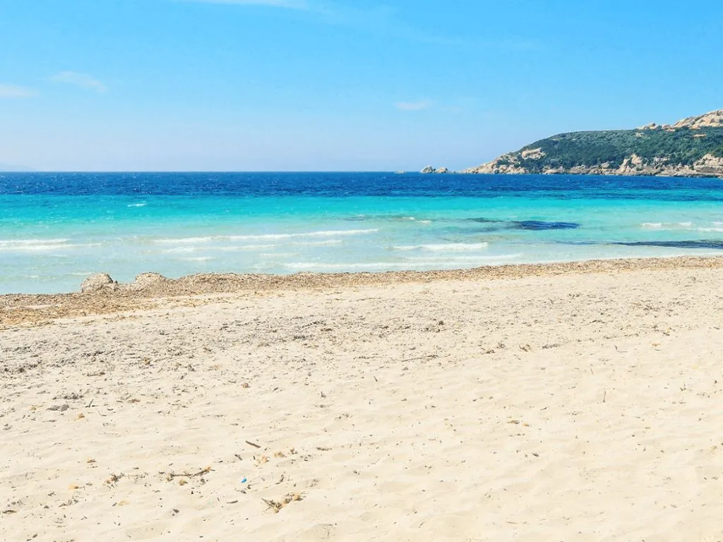 Spiaggia di sabbia chiara con mare turchese e promontorio roccioso sullo sfondo