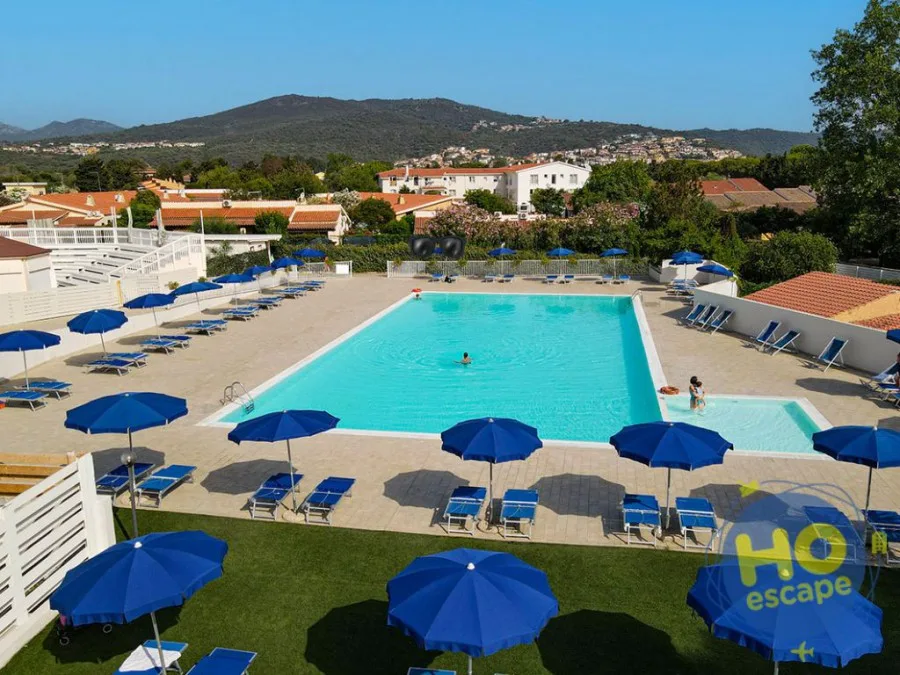 Ampia piscina con ombrelloni e lettini blu, pavimentazione chiara e colline verdi