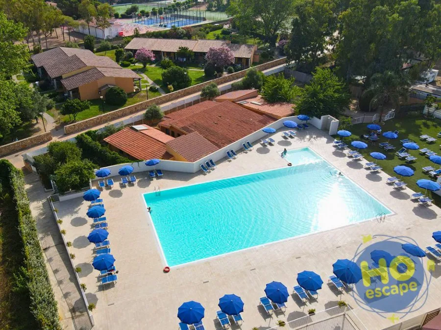 Vista aerea della grande piscina rettangolare con lettini e ombrelloni blu circondata da giardini e campi sportivi