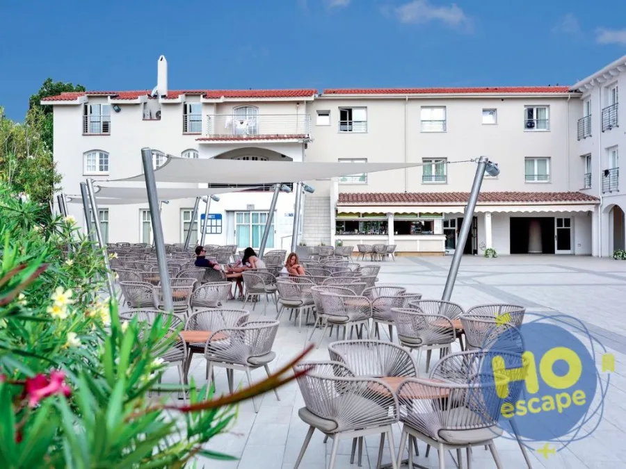 Terrazza esterna con tavoli e sedie in rattan grigio sotto teli ombreggianti, vista sull'edificio dell'hotel