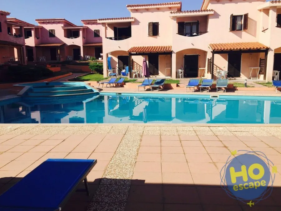 Piscina del residence con lettini blu e villette rosa in giornata di sole