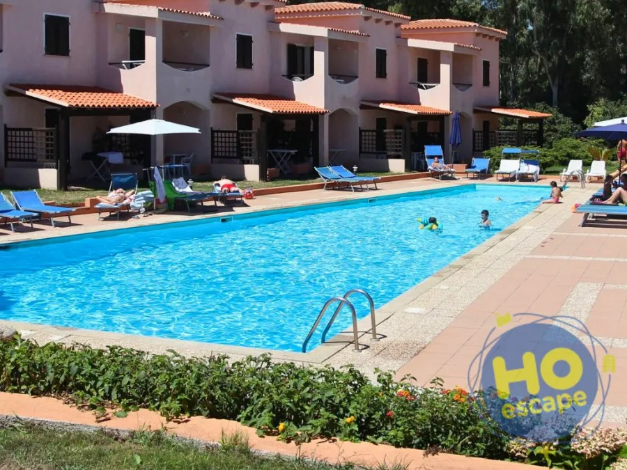 Piscina con bambini in acqua, lettini blu e villette beige circondate dal verde