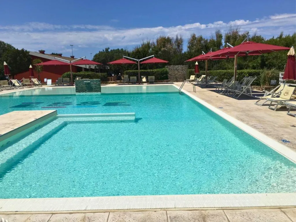 Piscina con acqua turchese, ombrelloni rossi e lettini su pavimentazione in pietra