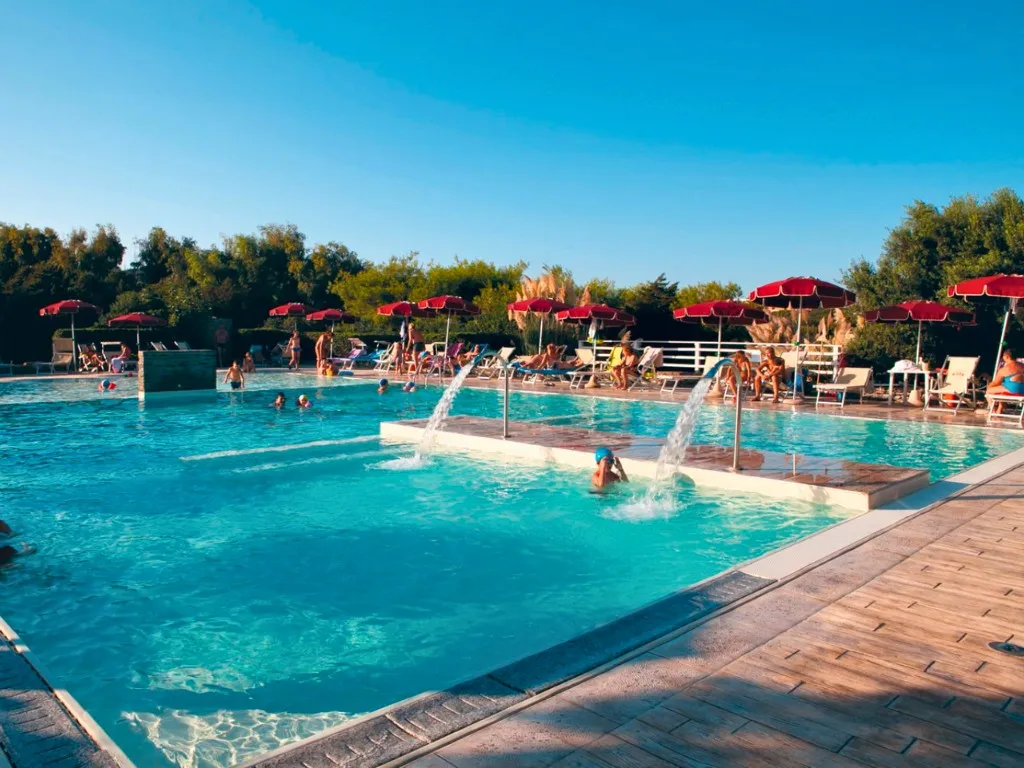 Piscina animata con bagnanti, giochi d'acqua e ombrelloni rossi al tramonto