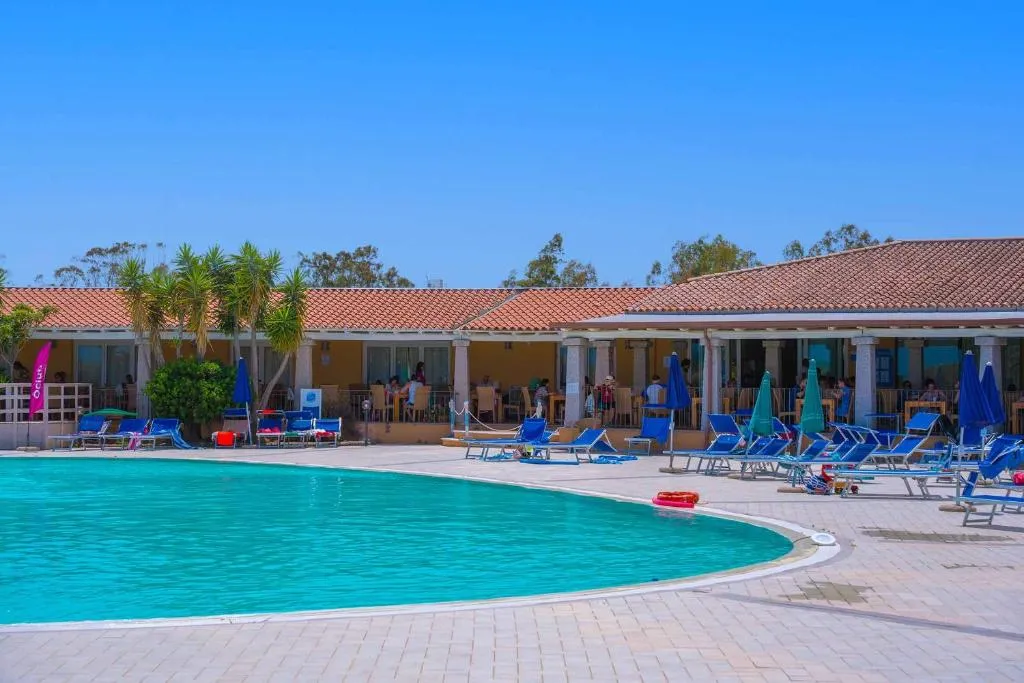 Piscina con lettini blu e ombrelloni davanti agli edifici del villaggio