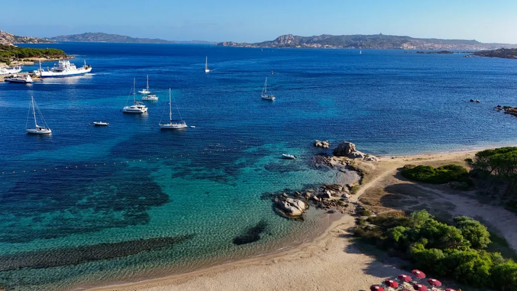 Spiaggia con mare turchese, barche a vela ormeggiate e rocce affioranti