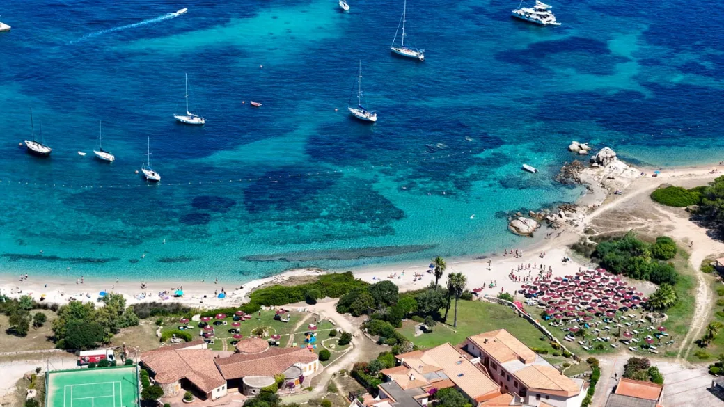 Spiaggia con ombrelloni rossi, barche a vela e mare turchese visti dall'alto