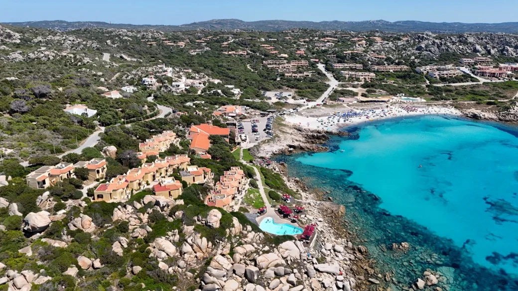 Veduta aerea del resort con piscina, cottage e spiaggia sulla costa rocciosa sarda