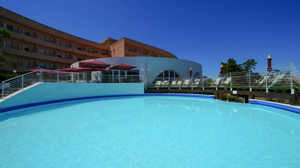 Piscina dell'hotel con lettini, ombrelloni rossi ed edificio curvo sullo sfondo