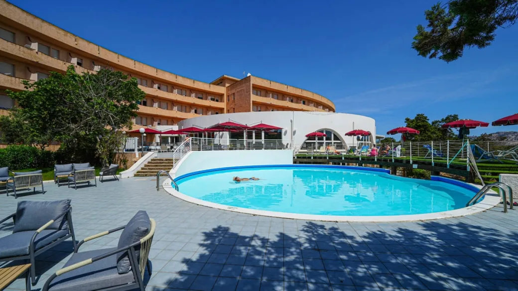 Piscina circolare con lettini e ombrelloni rossi davanti all'edificio curvo dell'hotel