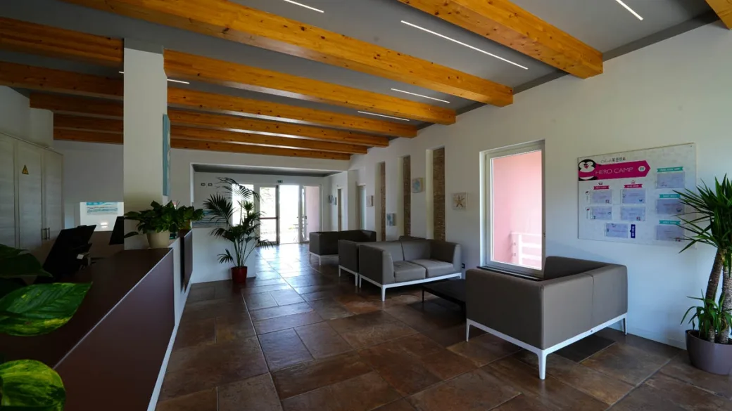 Area lounge con divani grigi, travi in legno e bacheca informativa