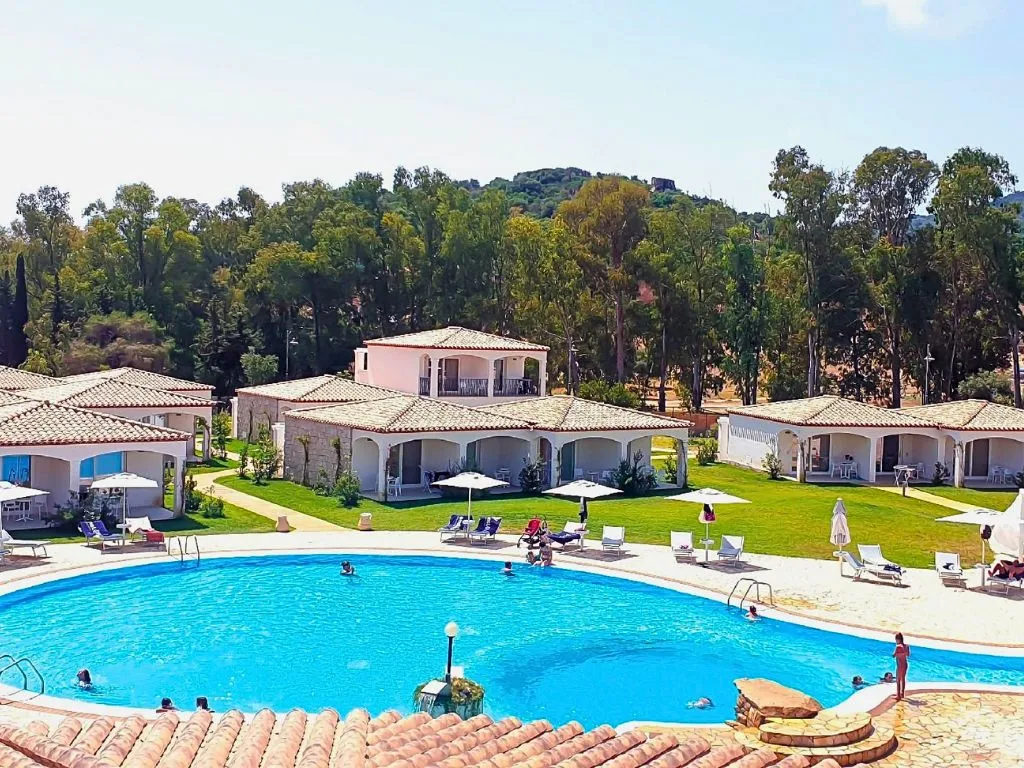 Veduta del resort con piscina, bagnanti e ville bianche con tetti in tegole immerse nel verde