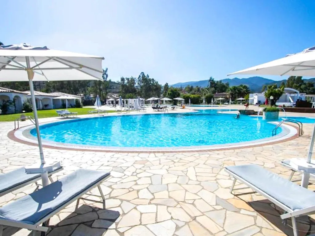 Piscina circolare con lettini, ombrelloni bianchi e pavimentazione in pietra con montagne sullo sfondo