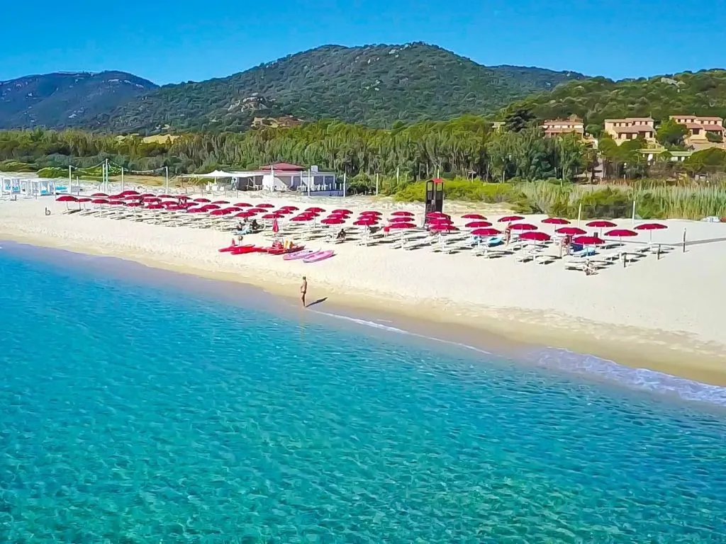 Spiaggia di sabbia con ombrelloni rossi e lettini davanti al mare turchese con colline verdi