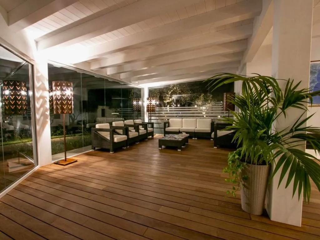 Area lounge coperta con divani bianchi, poltrone in rattan e pavimento in legno illuminata di sera