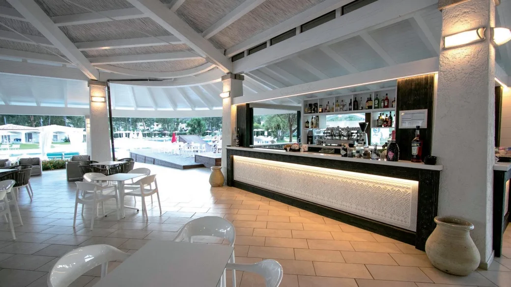 Bar interno del resort con bancone, tavolini bianchi e vista sulla piscina