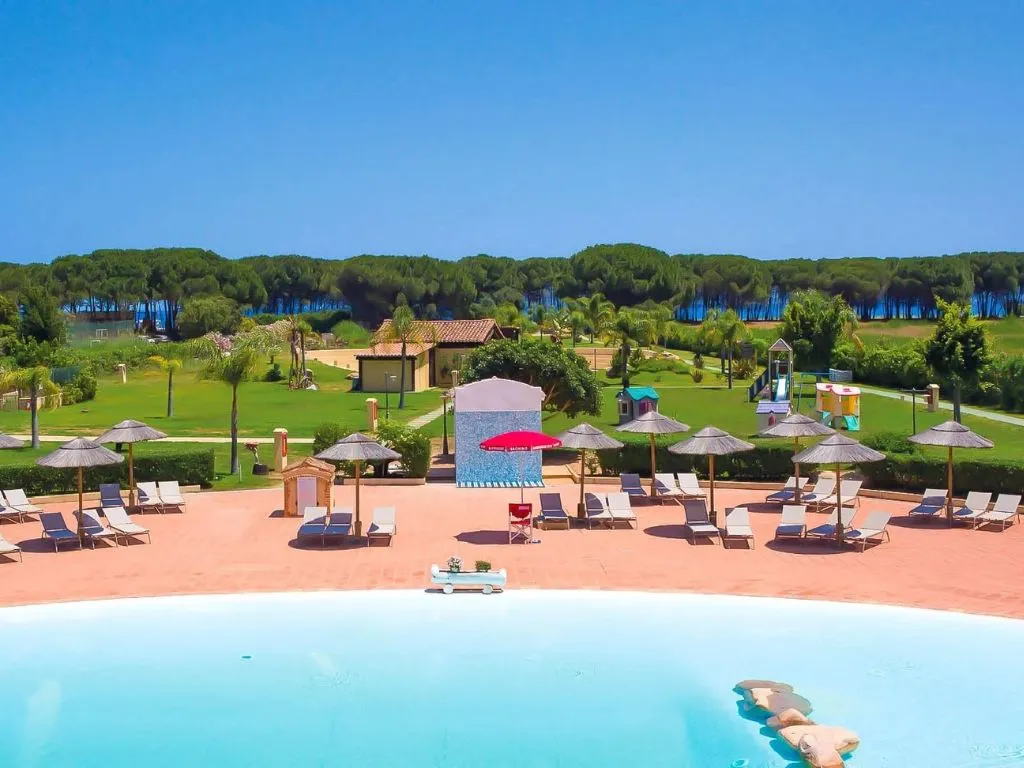 Piscina del resort con lettini, ombrelloni in paglia e ampio parco verde con area giochi