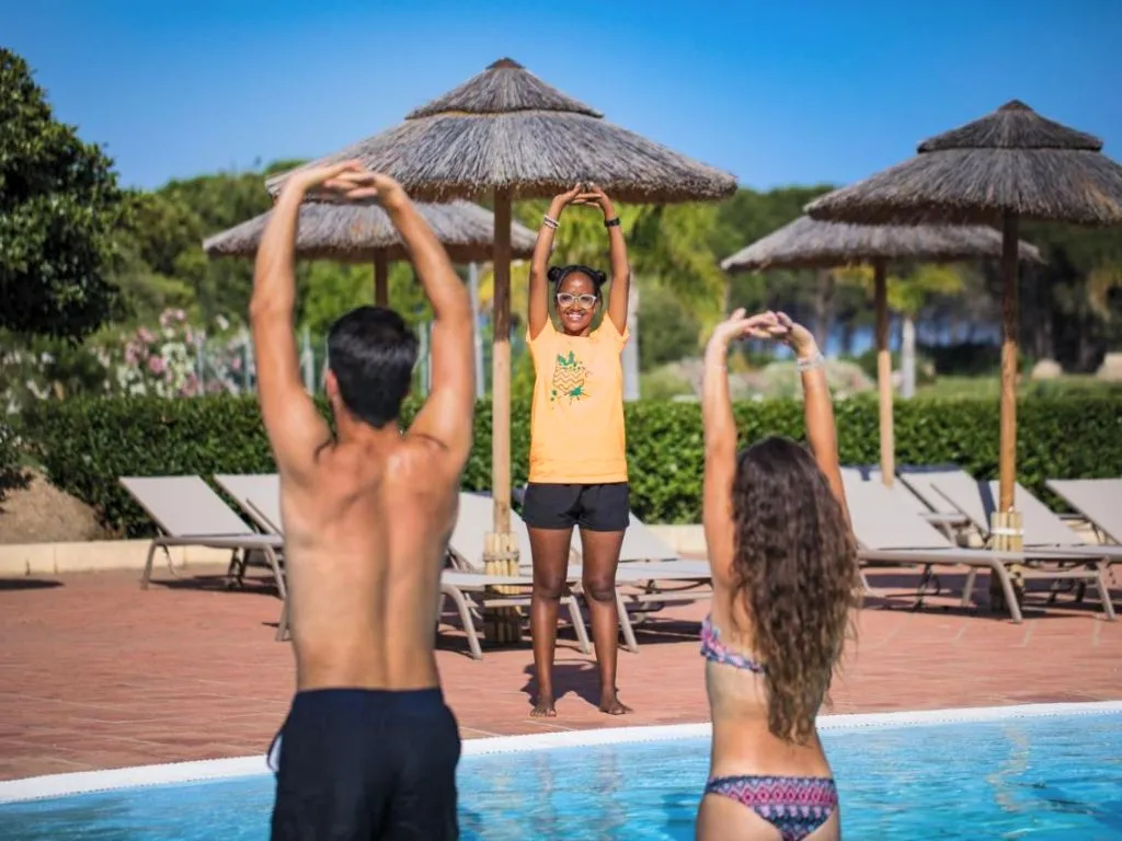 Attività di ginnastica a bordo piscina con animatrice e ospiti sotto ombrelloni in paglia