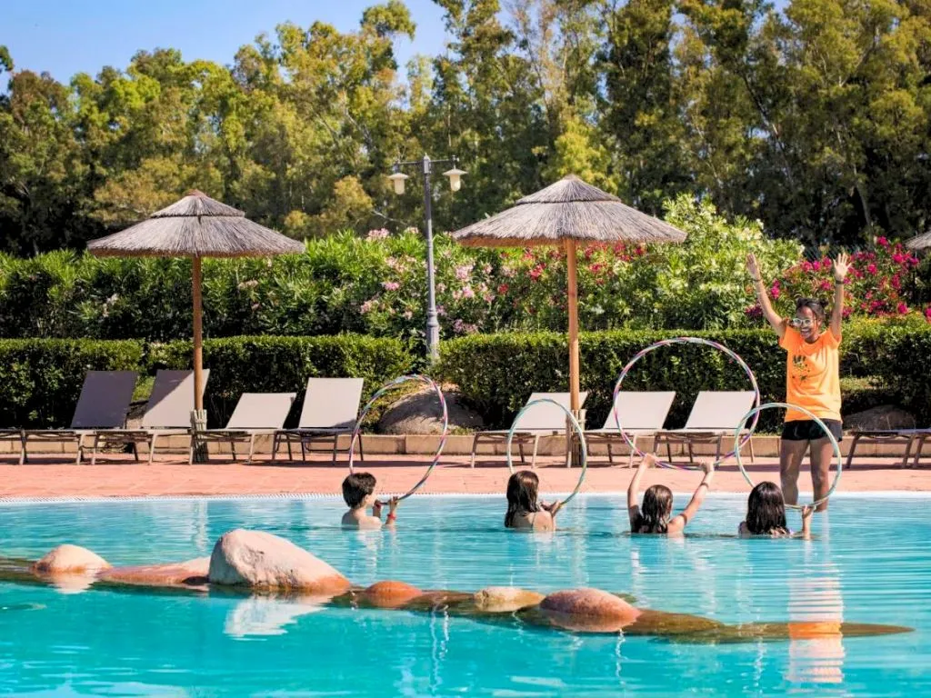 Animazione in piscina con bambini e istruttrice che giocano con cerchi tra rocce e ombrelloni