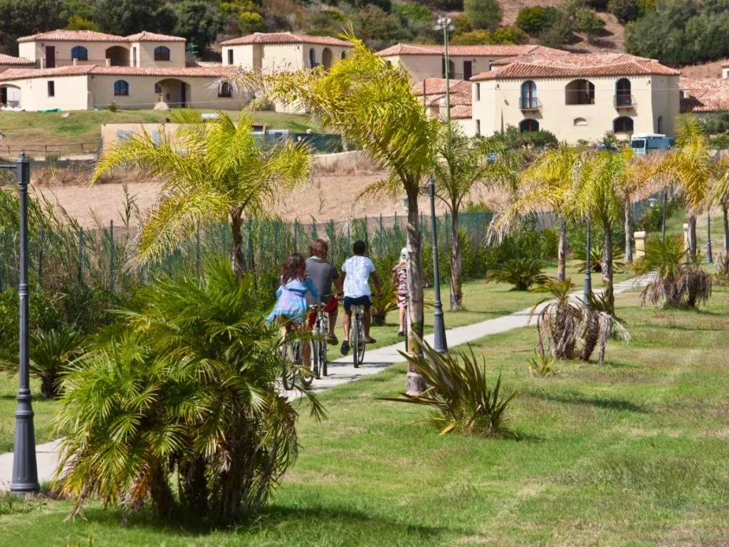 Ospiti in bicicletta lungo un viale alberato con palme e ville del resort sullo sfondo