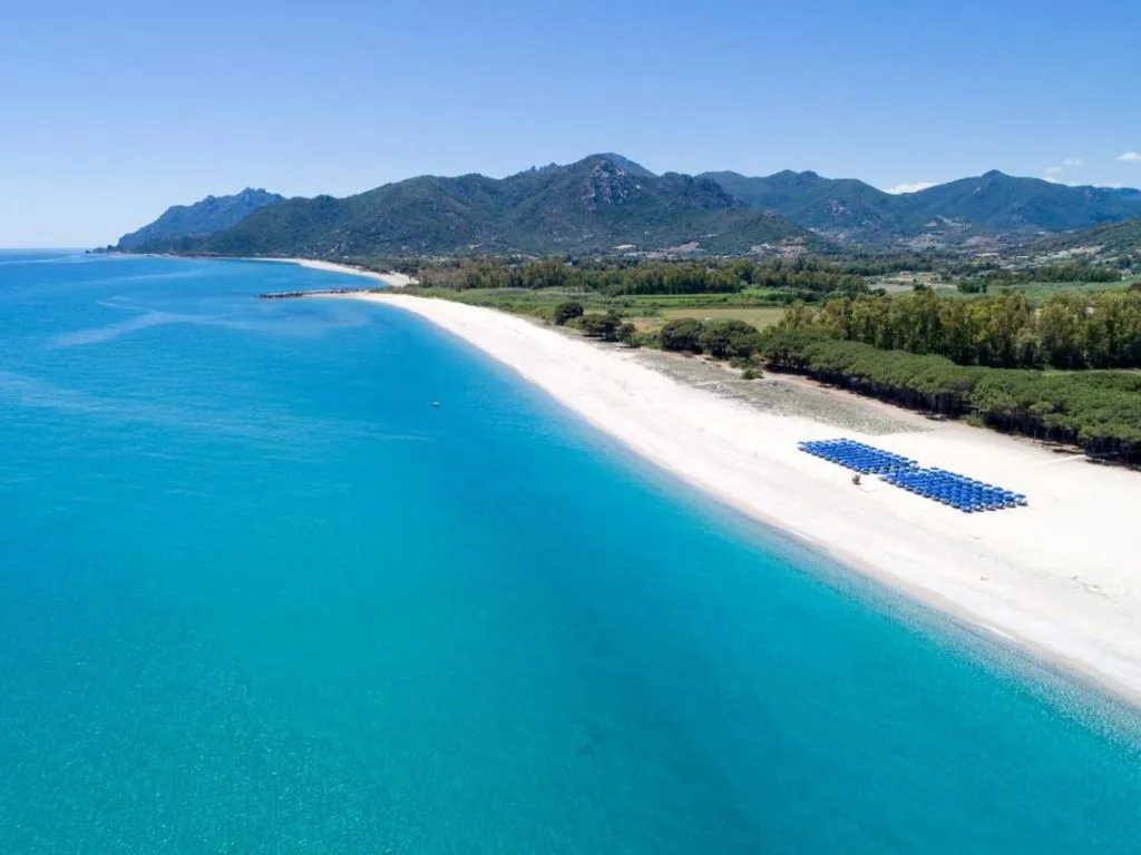 Veduta aerea della lunga spiaggia bianca con mare turchese, ombrelloni blu e montagne sullo sfondo