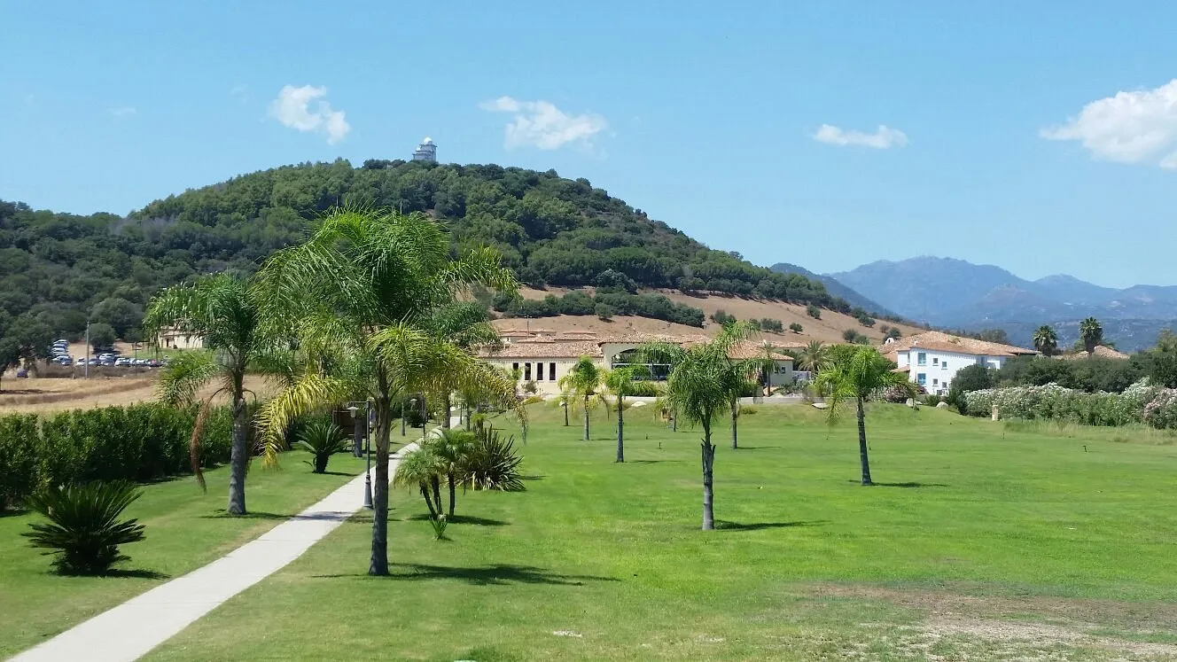 Vialetto tra prati verdi e palme con edificio del resort e collina boscosa sullo sfondo
