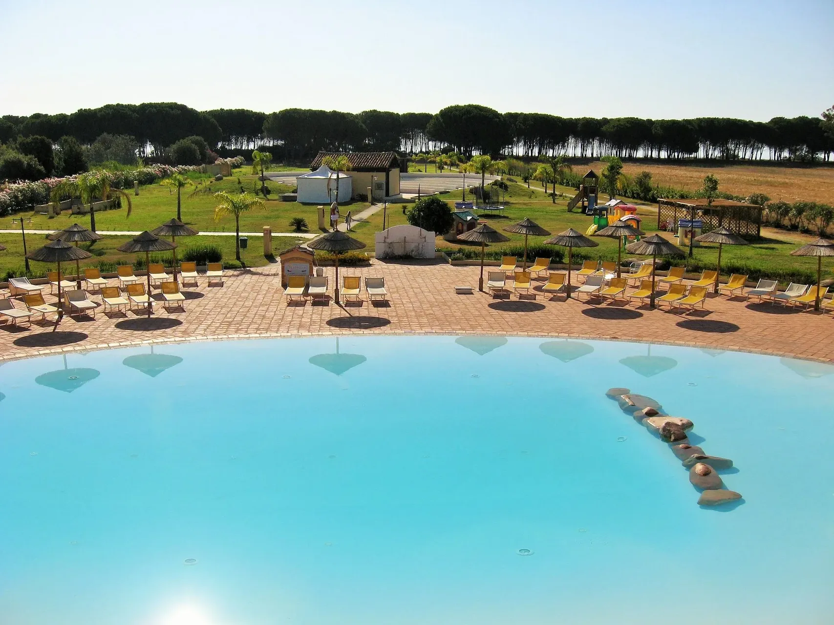 Grande piscina con lettini gialli, ombrelloni in paglia, prato e pineta sullo sfondo