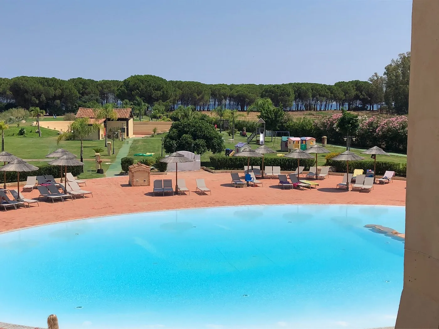 Piscina circondata da lettini e ombrelloni con parco verde e area giochi per bambini