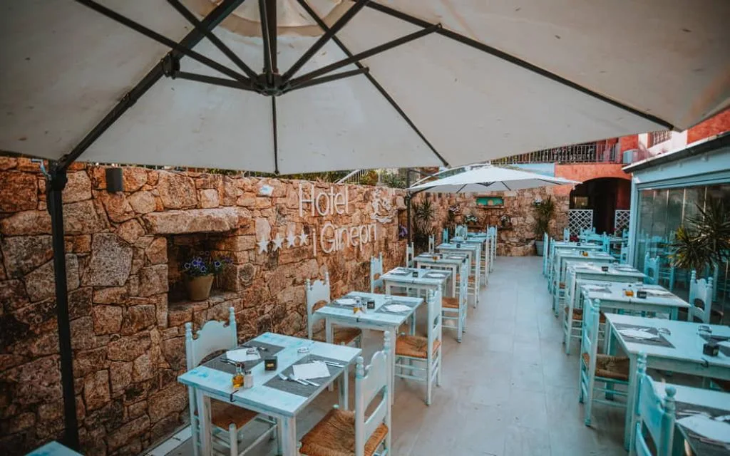Terrazza ristorante all'aperto con tavoli in legno bianco, sedie e parete in pietra sotto ombrelloni