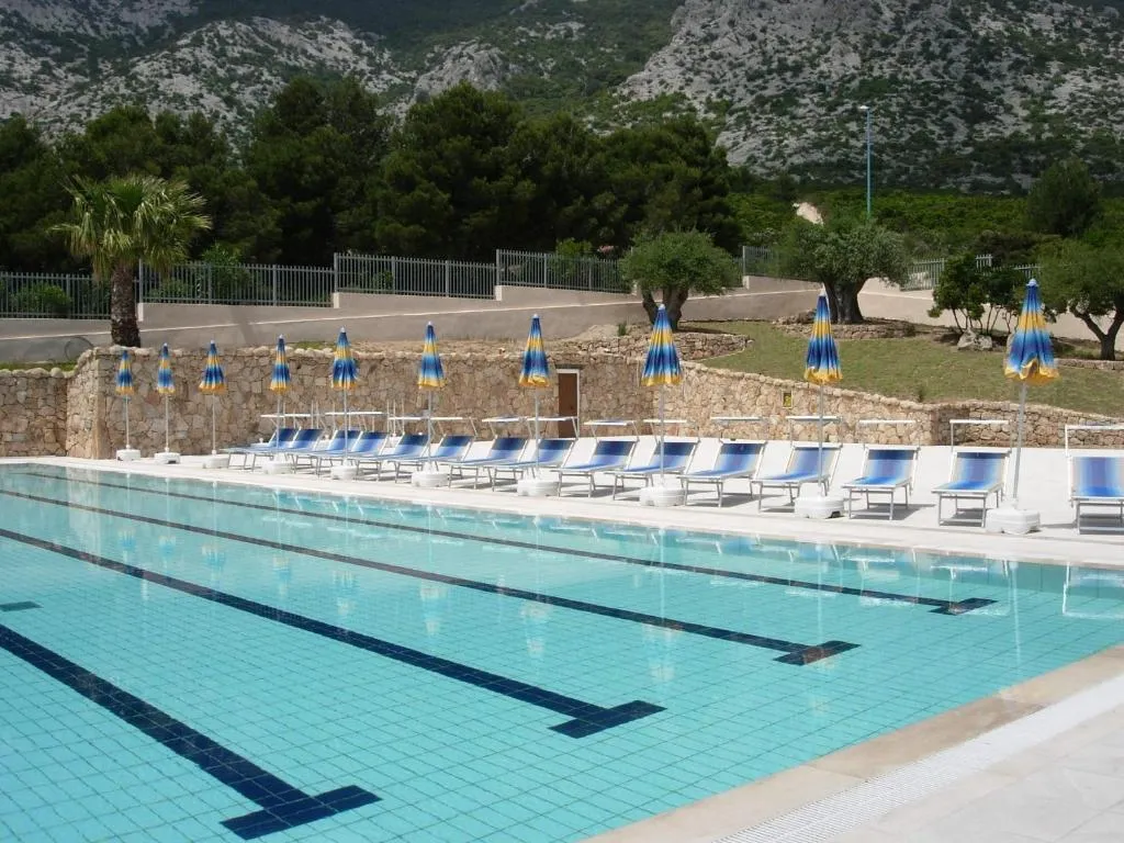 Piscina con lettini blu e ombrelloni a strisce davanti a montagne rocciose e pinete