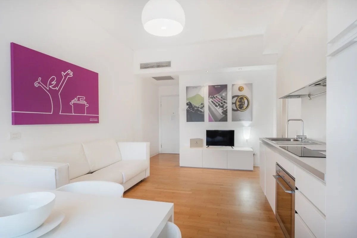 Soggiorno luminoso con divano bianco, quadro fucsia, TV e cucina attrezzata
