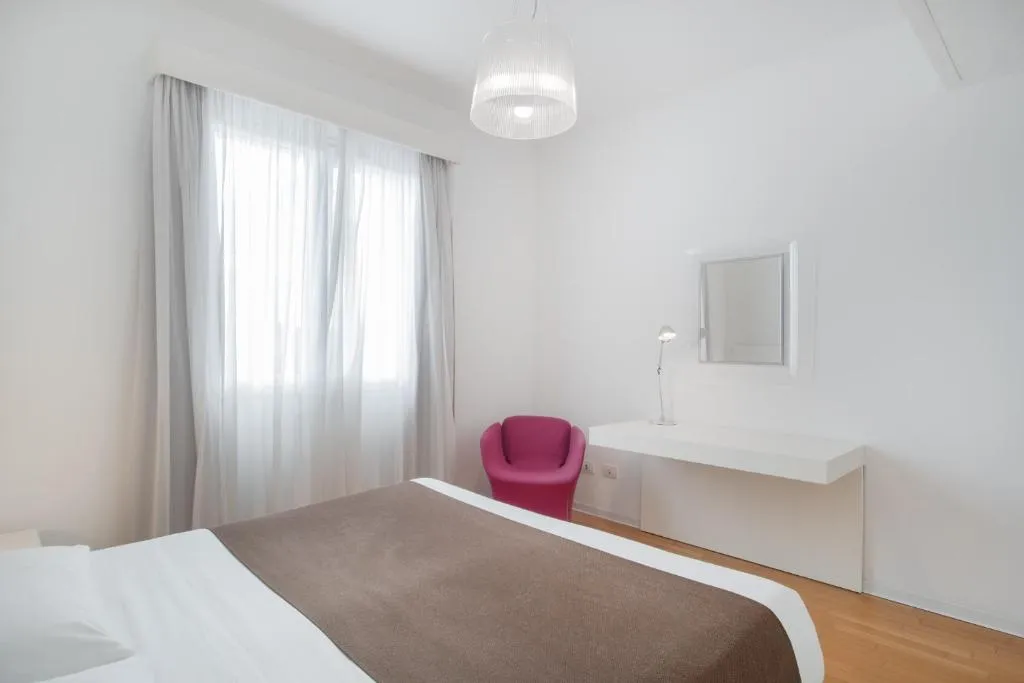 Camera da letto bianca con letto matrimoniale, scrivania e poltrona rosa accanto alla finestra