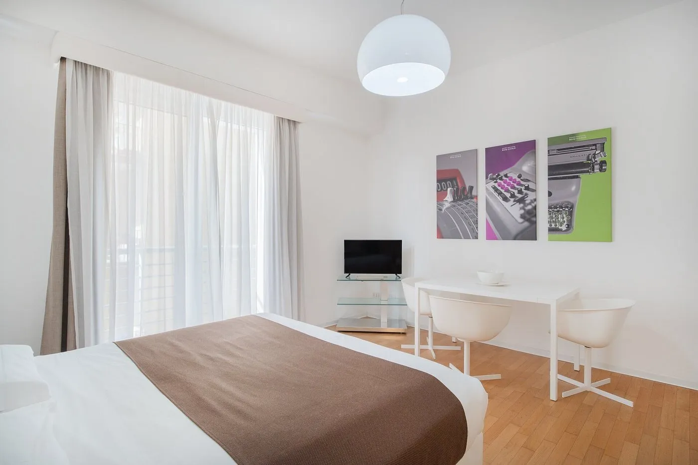 Camera con letto matrimoniale, tavolo da pranzo bianco, TV e quadri colorati a parete