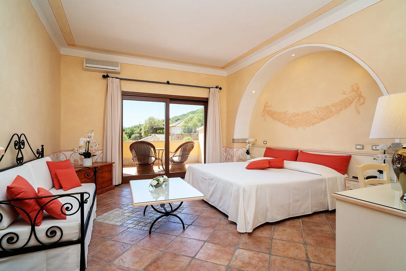 Camera con letto matrimoniale, divano con cuscini rossi e uscita sul balcone