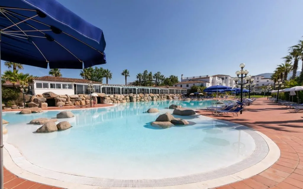 Ampia piscina laguna con rocce e bordo in cotto, palme e ombrelloni blu
