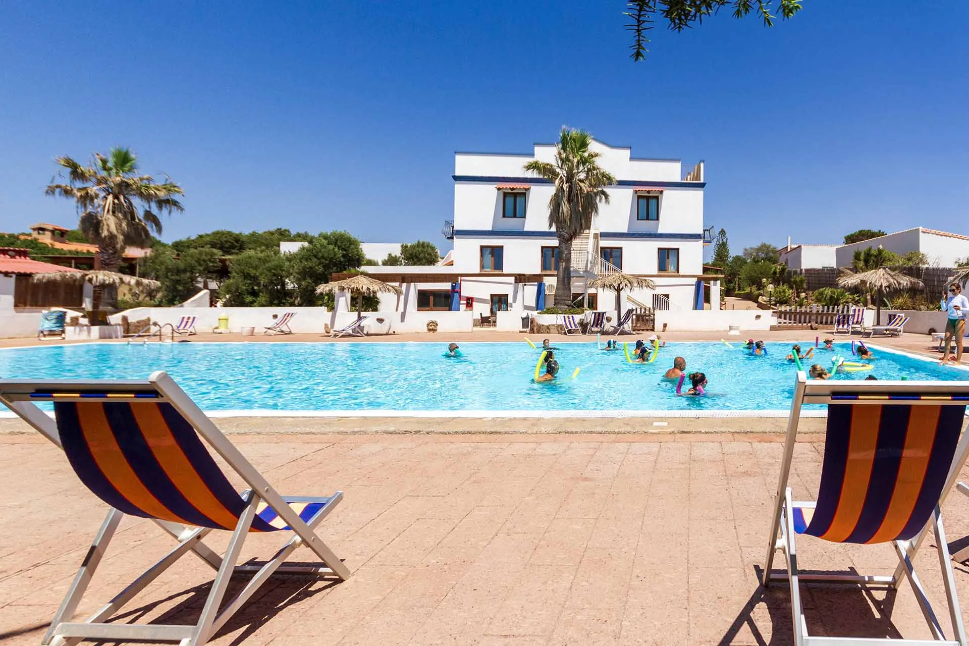 Piscina con lettini a righe e ospiti in acqua davanti all'hotel