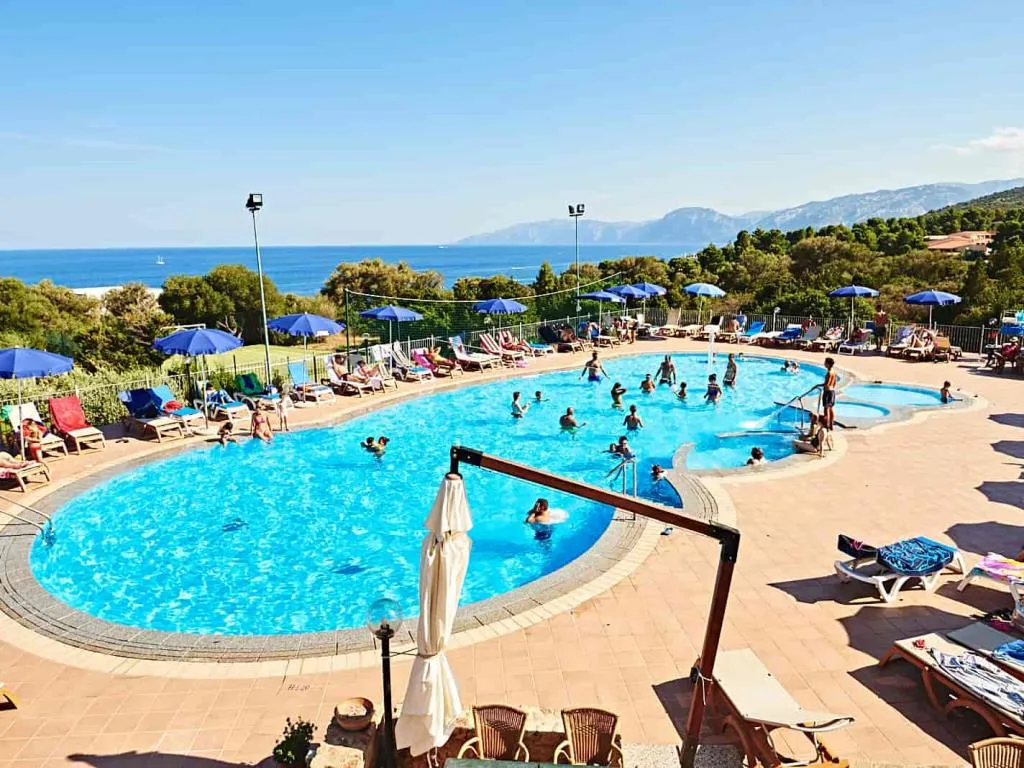 Grande piscina esterna con ospiti, ombrelloni blu e vista sul mare e le montagne