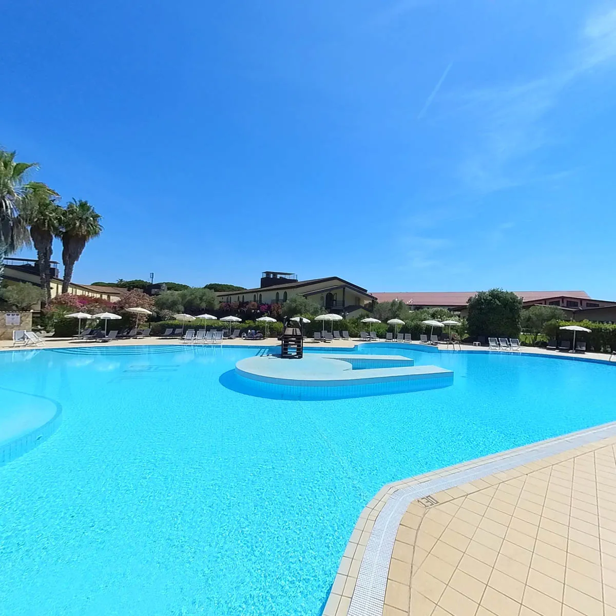 Piscina con lettini e ombrelloni, edifici dell'hotel e vegetazione sullo sfondo