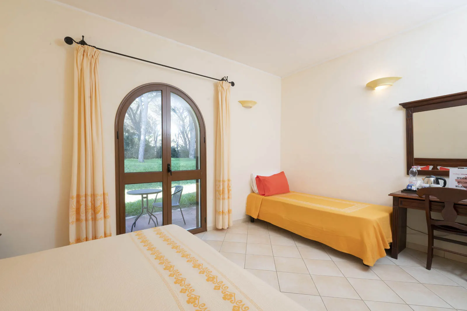 Camera con letto singolo, copriletto arancione e porta-finestra ad arco affacciata sul verde